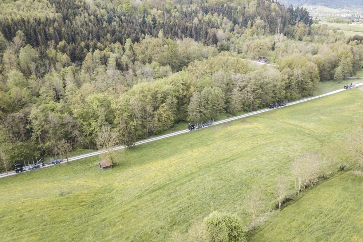 Drohnenaufnahme von 4 Rundholz-LKW's die auf einem staubigen Feldweg am Waldrand entlang fahren.