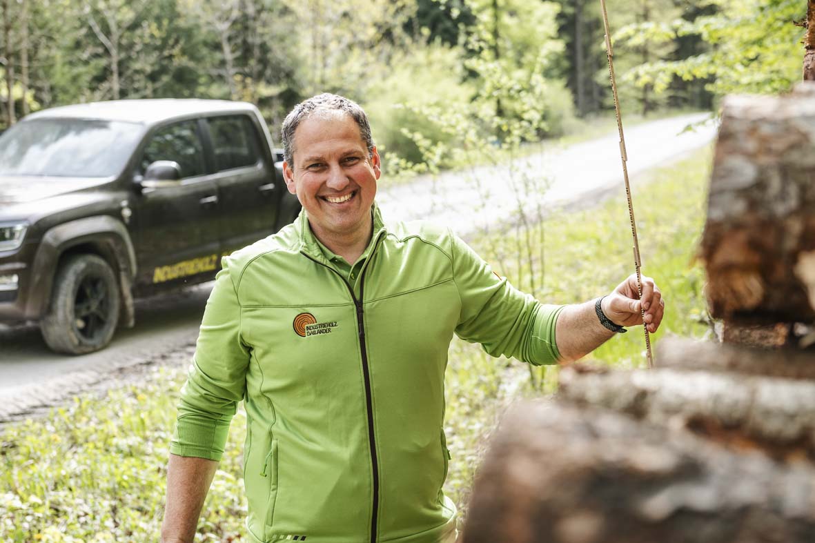 Markus Spieß im Wald bei der Übernahme von Holz. In der Hand hält er einen Meterstab und lächelt in die Kamera. Im Hintergrund ist ein brauner Pick Up zu sehen.