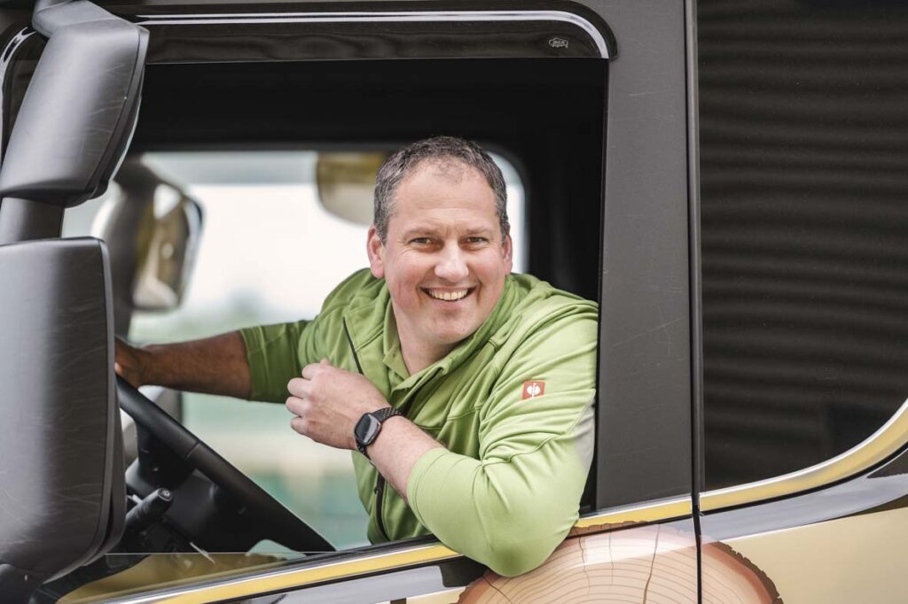 Markus Spieß fährt einen LKW und lehnt sich dabei aus dem Fenster. Er lächelt freundlich in die Kamera.