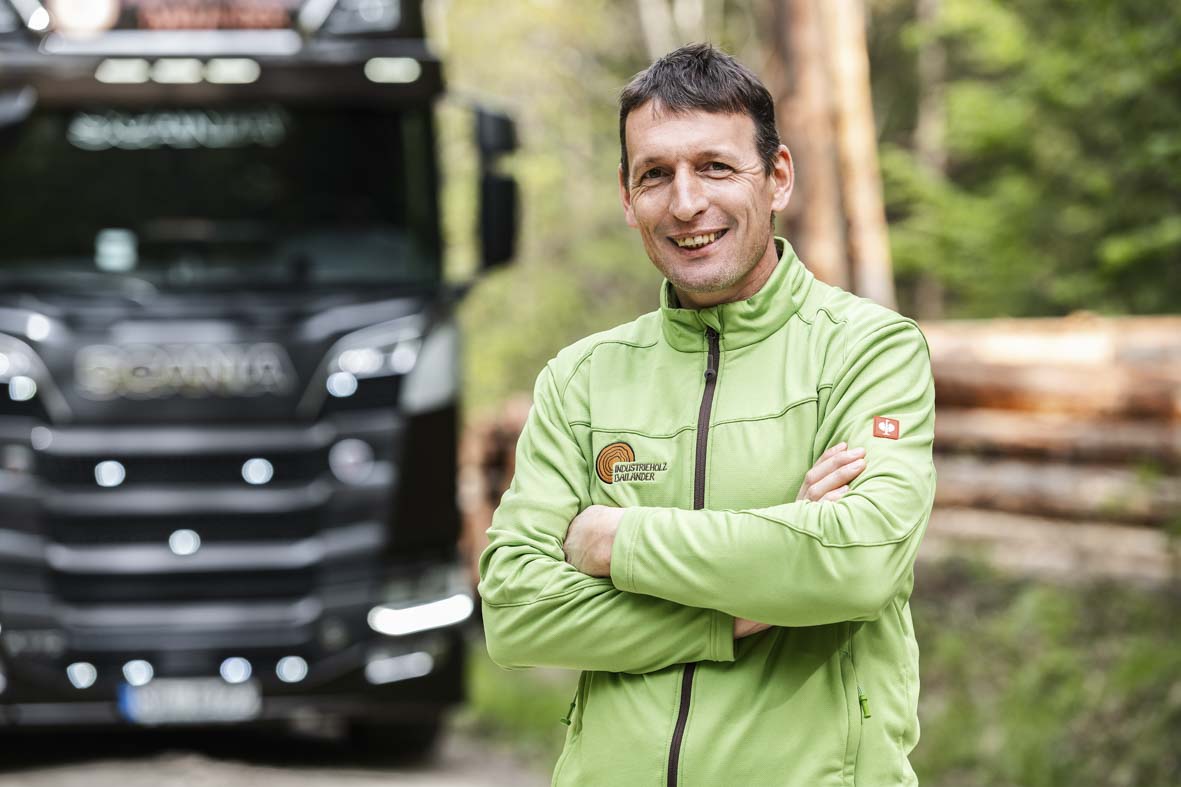 Portraitaufnahme im Wald von Andreas Grau. Im Hintergrund ein LKW und ein Stapel Baumstämme in der Unschärfe.