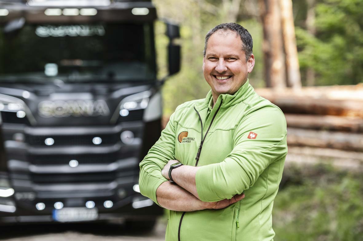 Portraitaufnahme im Wald von Markus Spieß. Im Hintergrund ein LKW und ein Stapel Baumstämme in der Unschärfe.