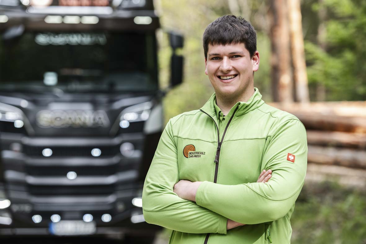 Portraitaufnahme im Wald von Marlon Spieß. Im Hintergrund ein LKW und ein Stapel Baumstämme in der Unschärfe.