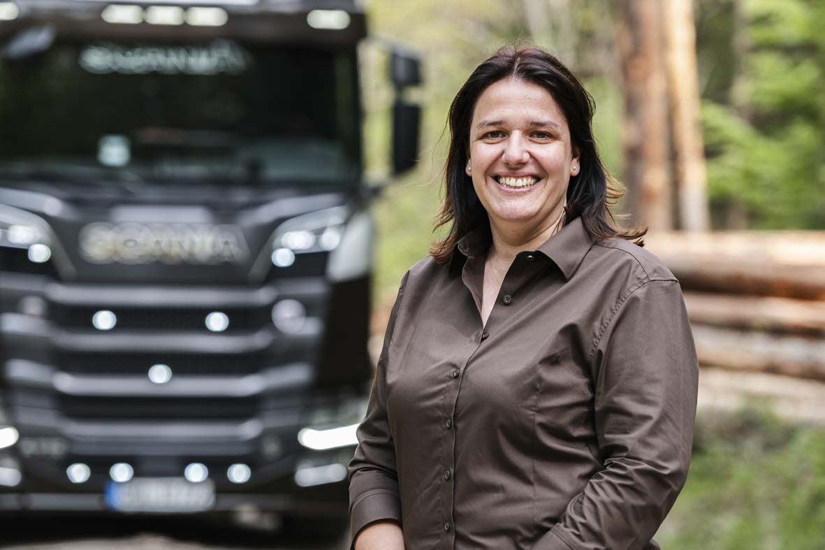 Portraitaufnahme im Wald von Melanie Spieß. Im Hintergrund ein LKW und ein Stapel Baumstämme in der Unschärfe.