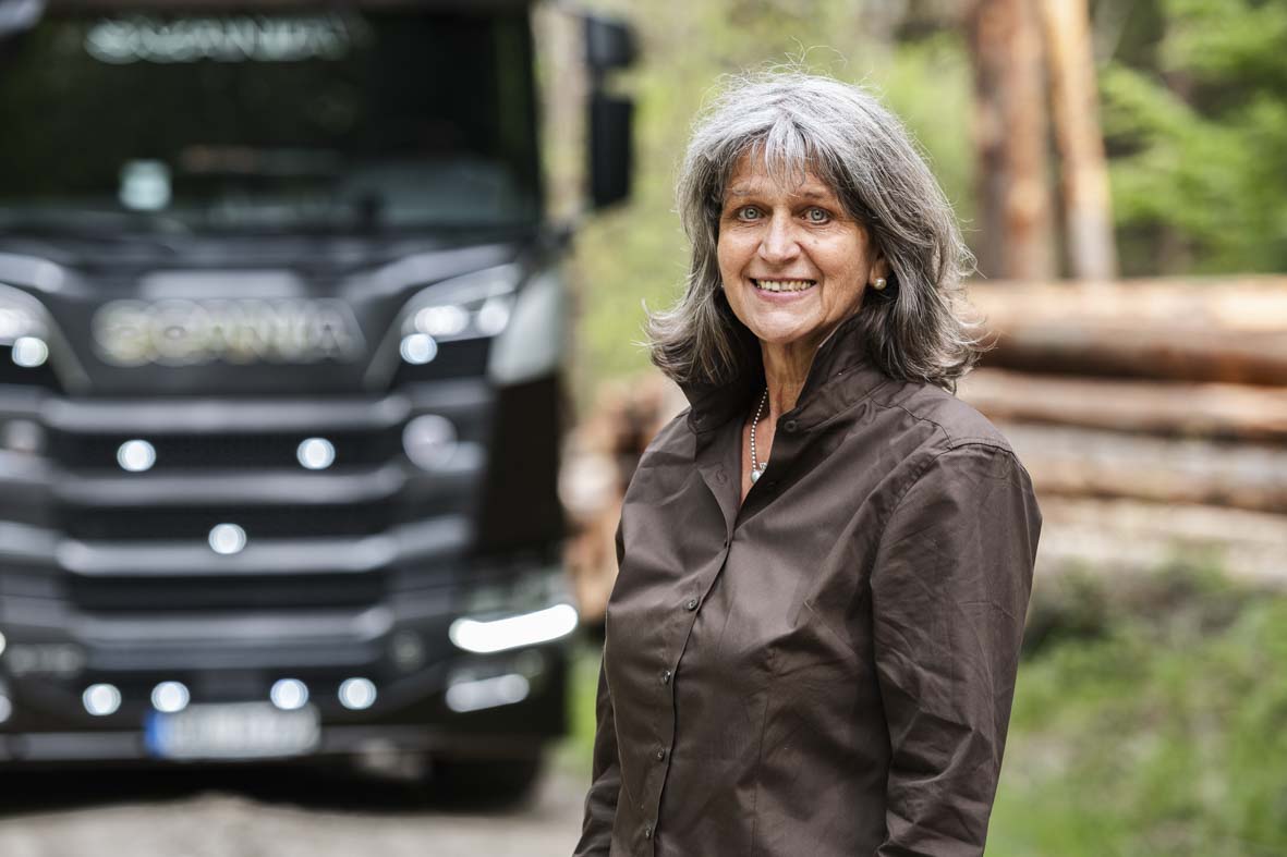 Portraitaufnahme im Wald von Mathilde Bailänder. Im Hintergrund ein LKW und ein Stapel Baumstämme in der Unschärfe.